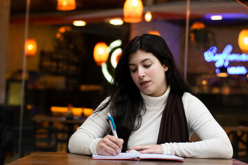 Seated woman writing. restaurant in the background. Thoughtful woman writing in a notebook. Woman sitting in a restaurant