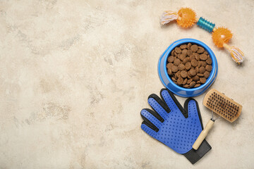 Bowl of dry pet food, toy and grooming brush on light background