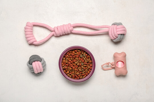 Bowl of dry pet food, toys and waste bags on light background