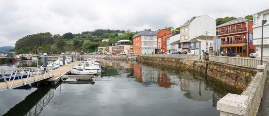 Fototapeta na wymiar O Porto do Barqueiro, La Coruña, pueblo marítimo en la costa gallega con edificios en la orilla reflejados en el agua del mar ,barcos amarrados y un bosque verde al fondo , en verano de 2021 España
