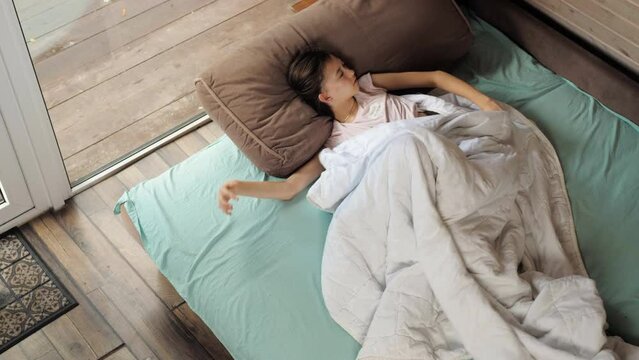 A teenage girl wakes up in a bed in a country house by a large window, pulls herself up and yawns.