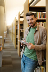 Positive smiling latin student standing in university library, using digital tablet, reading e books, education concept, modern technology