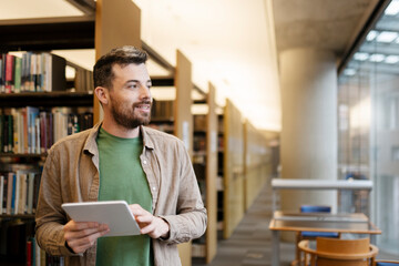 Smiling, handsome man stands in the university library, using digital tablet, looking away. Latin...