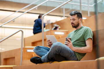 Young handsome latin man holding digital tablet reading ebook sitting on steps of university campus