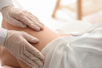 Dermatologist examining moles on young woman's leg in clinic, closeup