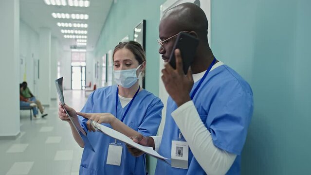 African American practitioner standing in hospital corridor talking on phone, his Caucasian female colleague interrupting him to ask question about X-ray shot
