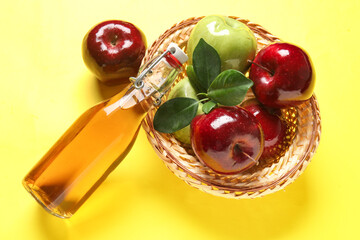 Glass bottle of fresh apple cider vinegar and basket with fruits on yellow background
