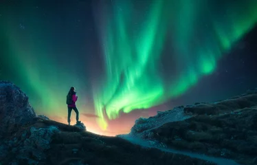Poster Northern lights and young woman on mountain peak at night. Aurora borealis and silhouette of alone girl on mountain trail. Landscape with polar lights. Starry sky with bright aurora. Travel background © den-belitsky