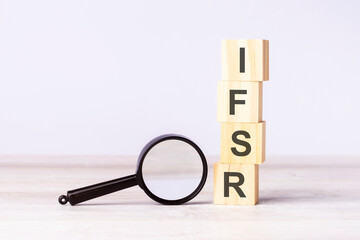 magnifying glass and wooden blocks with text IFSR on wood table, grey background