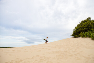 Man with a sand board wearing a straw hat walking up a sand dune in Balneario Atlantico beach, Arroio do Sal, RS, Brazil