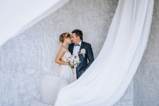 A young groom and a beautiful bride model blonde in a white dress with a bouquet in their hands are hugging in a long fabric, curtains in the interior. Wedding photography, portrait of the newlyweds.