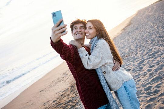 Beautiful young couple making photos with mobile phone in a cold winter on the beach.