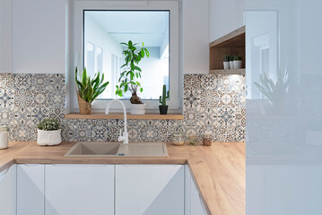 Kitchen sink near window in modern interior. Tiles pattern on the wall, wooden counter top in...