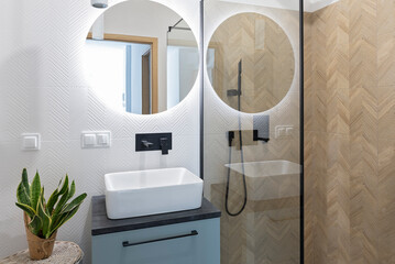 Ceramic washbasin with tap, wooden tiles in the shower with glass and round mirror with led lights on the wall.Interior of bathroom.