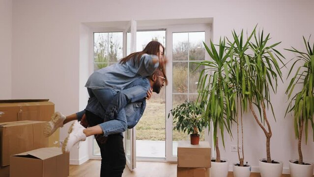 Happy african american man and caucasian woman are fooling around during moving to new house. The woman jumped on the man's back in a game of piggyback or horsey. The room is furnished with boxes