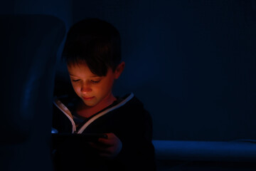Little boy playing with tablet or cellphone in the dark. Kids face illuminated by the light of the screen. Gadget addicted children. Prolonged telephone play negatively affects eye sight and health