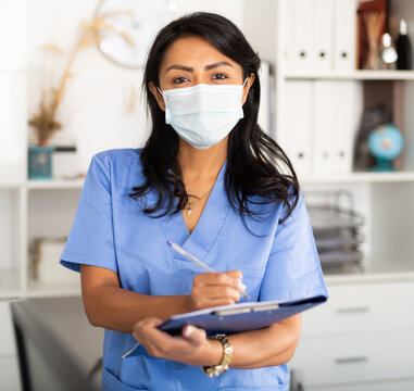 Hispanic woman general practitioner in uniform and face mask standing in clinic, filling clipboard with medical records.
