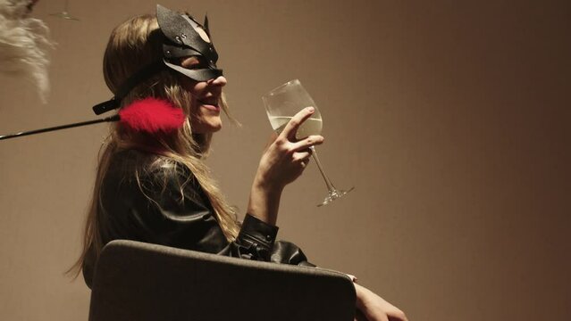A woman in a black masquerade mask drinks alcohol from a glass and other woman playing with her with a feather tickler