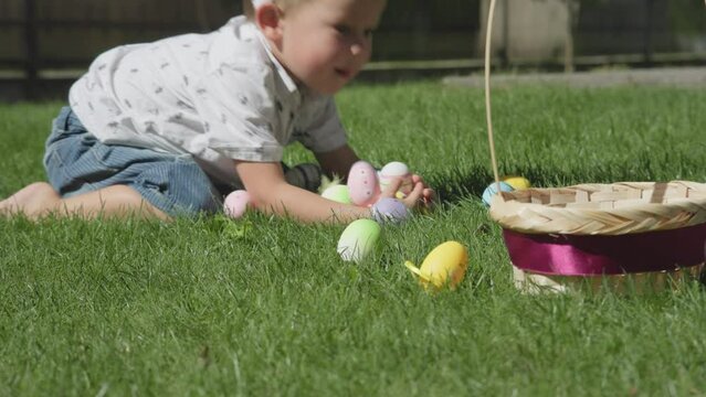 happy child kid boy playing with colored Easter eggs on green spring grass smiling laughing grab and fall Easter eggs. Preschooler boy wearing Easter bunny ears white shirt blue jeans rolling over 