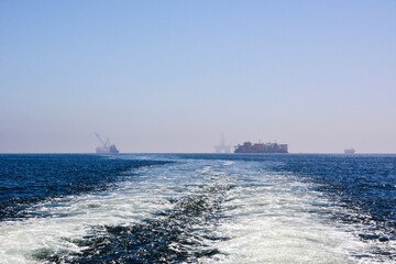 Off in the sea are oil and gas drilling platforms. In the foreground is a wake of waves from a ship