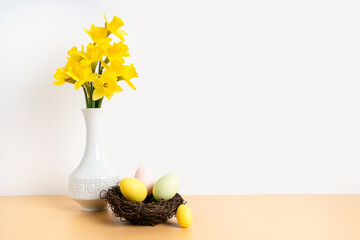 Colorful pastel easter eggs in nest and bouquet of daffodil flowers in white vase on beige table and white background. Happy Easter and holiday preparation. Festive decoration
