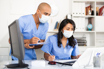 Professional doctor in surgical mask working in office with female assistant, filling up medical forms in laptop - obrazy, fototapety, plakaty