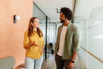 Young enthusiastic female colleague explains new strategy to male employee and gesturing. Indoor portrait of diverse work team discussing, walking through hallway in modern coworking office space
