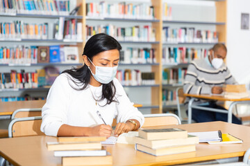 Young adult woman in protective face mask working with books, finding information at library