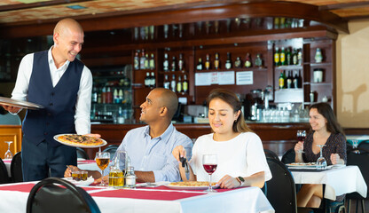 Fototapeta na wymiar Portrait of smiling polite waiter serving delicious pizza to positive hispanic couple in restaurant