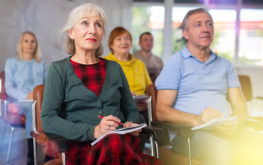 Focused motivated older woman listening to lecture in auditorium with group. Seniors education concept