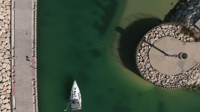 Aerial view of a Small sail boat leaving a marina slowly passing by a lighthouse