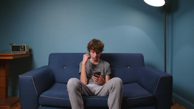 Teenager sits on the couch without talking. Boy addicted to gadgets, using mobile phone, laptop at home. Internet obsession concept
