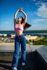 Beautiful sexy slim brunette caucasian girl in pink top and blue jeans standing on the roof attractively posing with softly blurred sky background