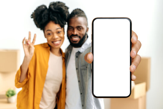 African american couple in defocus, newlyweds, show smart phone with empty white mock-up screen while stand in living room against the background of cardboard boxes with things in their new home