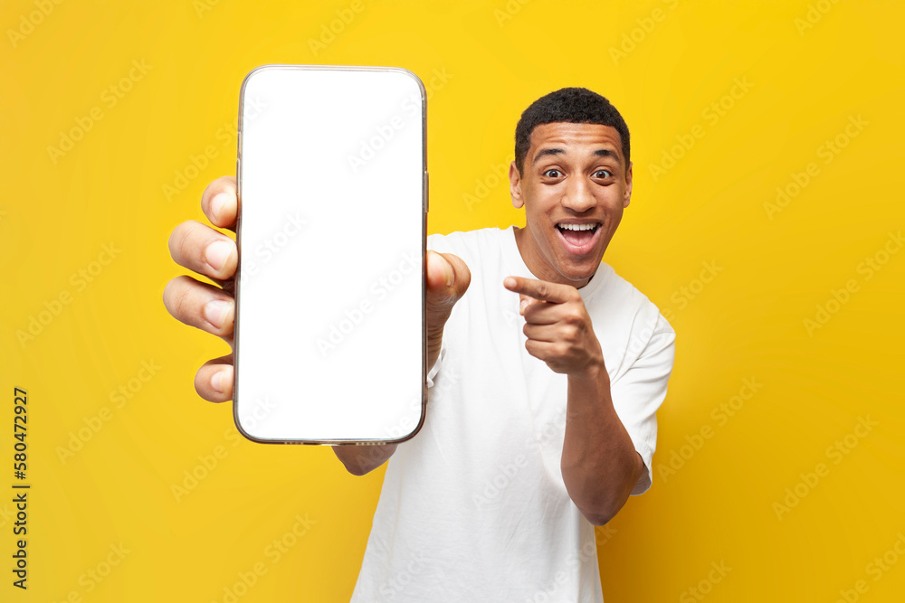 Wall mural young african american guy in white t-shirt shows blank screen of smartphone on yellow isolated background