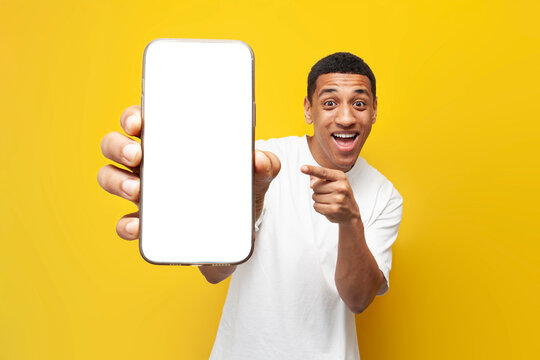 young african american guy in white t-shirt shows blank screen of smartphone on yellow isolated background