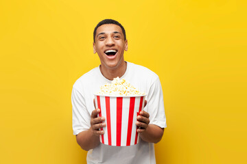 young african american guy with popcorn watching comedy and laughing on yellow isolated background,...