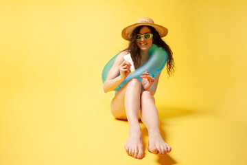 young girl in blue swimsuit with swimming inflatable ring sits on the beach and holds sunscreen on yellow background