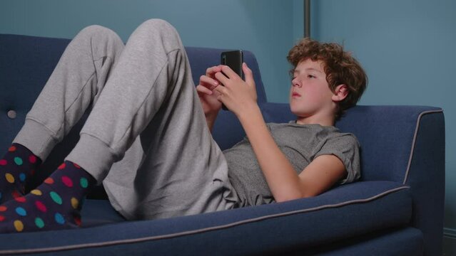 Teenager sits on the couch without talking. Boy addicted to gadgets, using mobile phone, laptop at home. Internet obsession concept