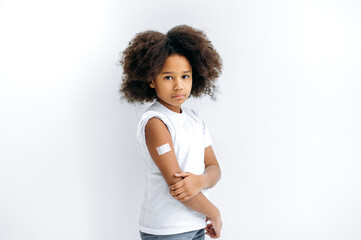 Upset African American curly haired little girl, with a band-aid on her shoulder after vaccination, stand on isolated white background, looks at camera. Virus protection, immunity concept