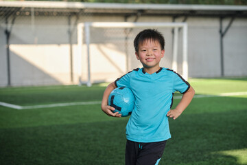 Cute little smiling Asian 7 years old child holding a soccer ball, football player in soccer uniform is playing football at Training Session, Soccer Drills for Kids, Selective focus - obrazy, fototapety, plakaty