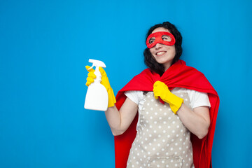 young girl cleaner in gloves and superman costume holds detergent on blue background