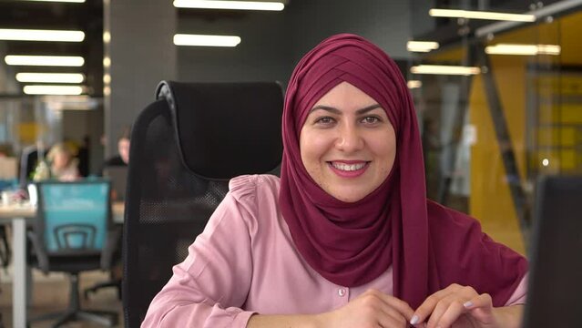 A happy Muslim woman in the office at her desk and laptop smiling looks at the camera. Multinational team