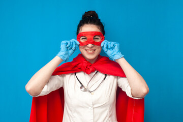 girl doctor in uniform in superman costume on blue background, female nurse puts on superhero mask, healthcare concept - obrazy, fototapety, plakaty