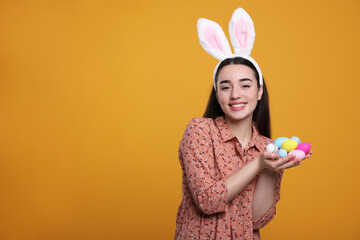 Happy woman in bunny ears headband holding painted Easter eggs on orange background. Space for text.