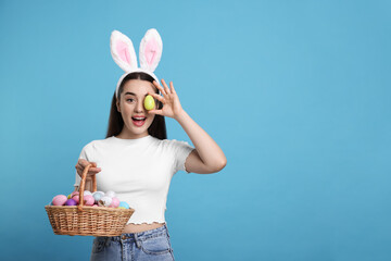 Happy woman in bunny ears headband holding wicker basket of painted Easter eggs on turquoise...