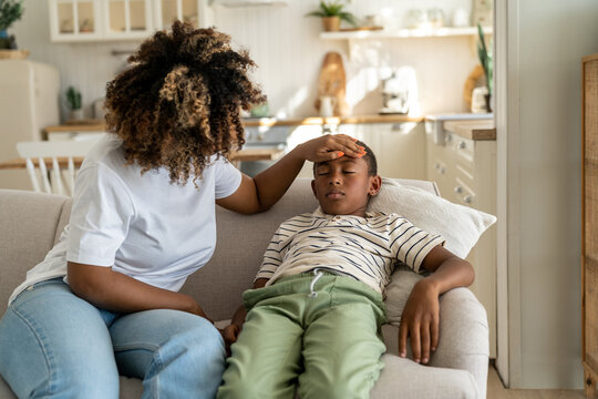 Fever In Kids. African American Mother Touching Forehead Of Sick Little Boy Son At Home, Caring Parent Mom Checking Childs Temperature With Hand. Schoolboy Lying On Sofa Feeling Unwell And Ill