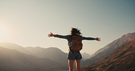 Young girl standing on top of mountain and victoriously raising hands up, looking far away -...