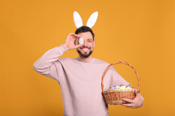 Happy man in bunny ears headband holding wicker basket with painted Easter eggs on orange background