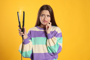 Upset young woman with flattening iron on yellow background. Hair damage